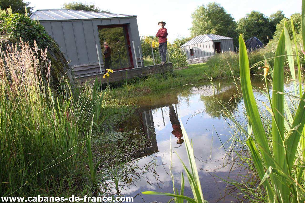 2172 Cabane moderne au bord dun étang, entourée de verdure luxuriante.