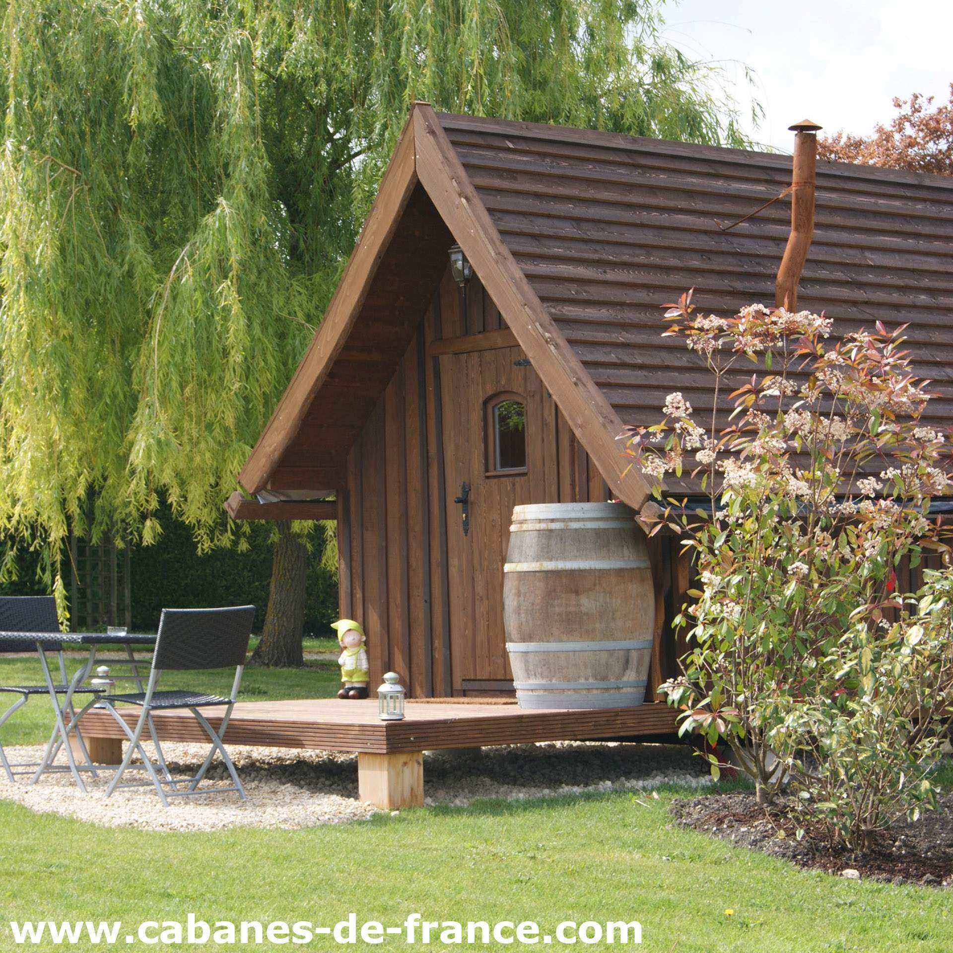 Charmante cabane en bois avec terrasse et baril décoratif dans un jardin verdoyant.