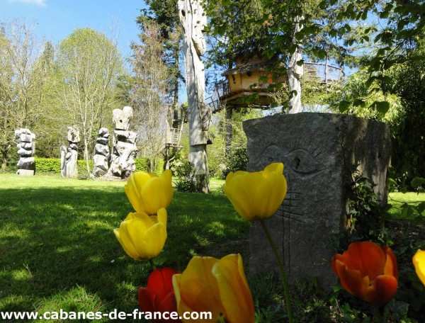 Cabane dans les arbres entourée de sculptures et de fleurs colorées.