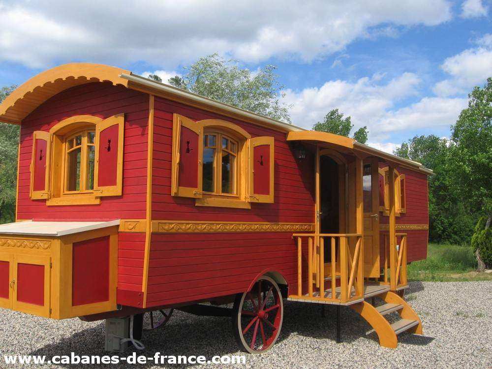 Charmante roulotte rouge avec fenêtres jaunes, entourée de verdure.