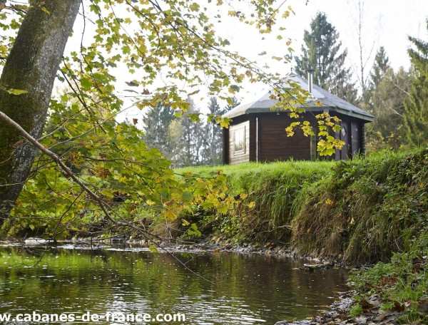 Cabane en bois au bord dun ruisseau, entourée darbres verdoyants.