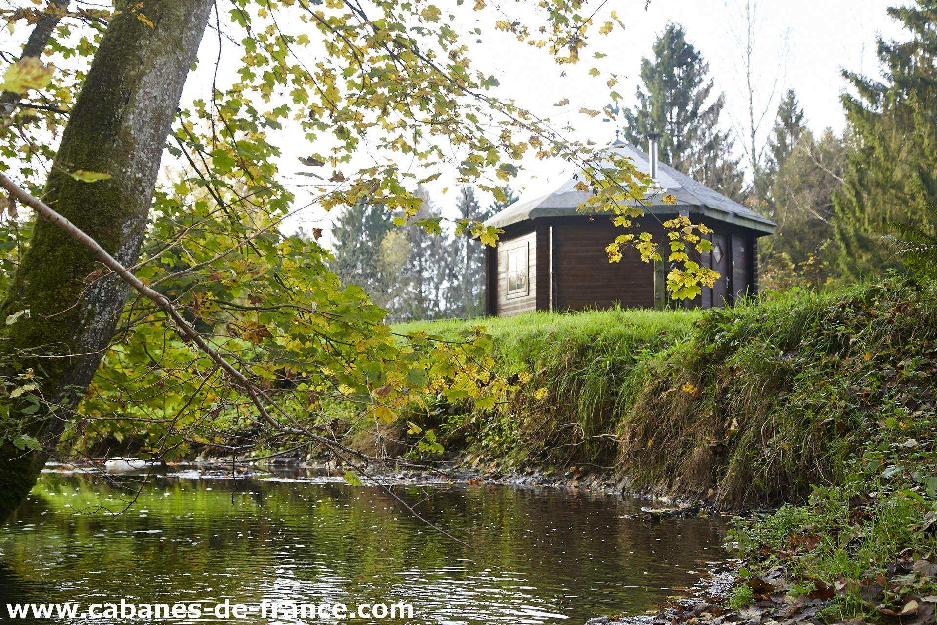 Cabane en bois au bord dun ruisseau, entourée darbres verdoyants.