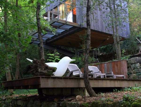 Cabane moderne suspendue, entourée darbres, avec terrasse en bois et mobilier design.