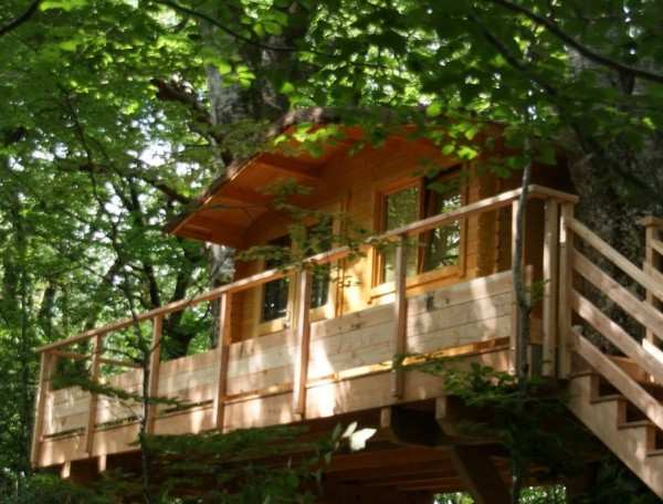 Cabane dans les arbres, en bois, avec balcon surplombant la forêt verdoyante.