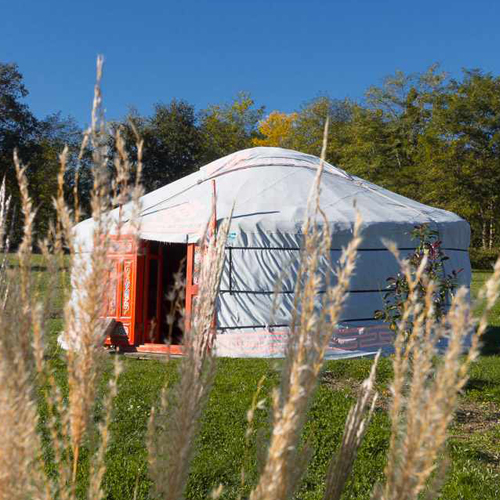 CC-Nomade-photo5 Yourte spacieuse entourée de verdure, avec une porte en bois colorée.