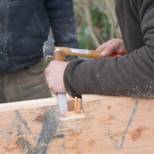 Hébergement en bois, artisan au travail sur une pièce de bois, ambiance chaleureuse.