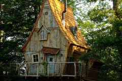 Cabane dans les arbres en bois, avec un toit en ardoise et entourée de verdure.