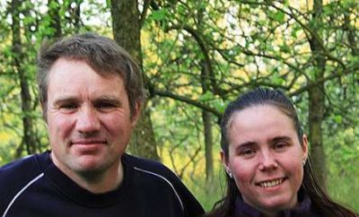 Couple souriant devant un gîte rural, entouré darbres verdoyants.