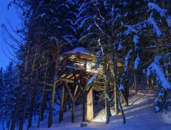 Cabane perchée dans les arbres, illuminée, entourée de neige et de sapins.