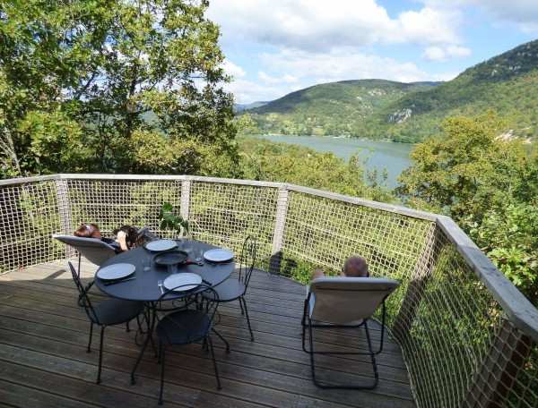 Chalet en bois avec terrasse panoramique sur un lac et des montagnes verdoyantes.