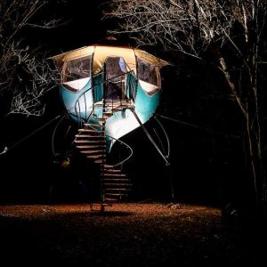 Cabane perchée illuminée, accessible par un escalier en colimaçon, au milieu des arbres.