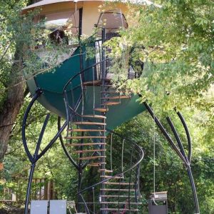 Cabane dans les arbres avec une terrasse, accessible par un escalier spiral.