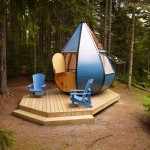 Cabane moderne en forme de goutte, entourée darbres, avec chaises bleues sur la terrasse.