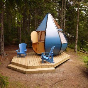 Cabane moderne en forme de goutte, entourée darbres, avec chaises bleues sur la terrasse.