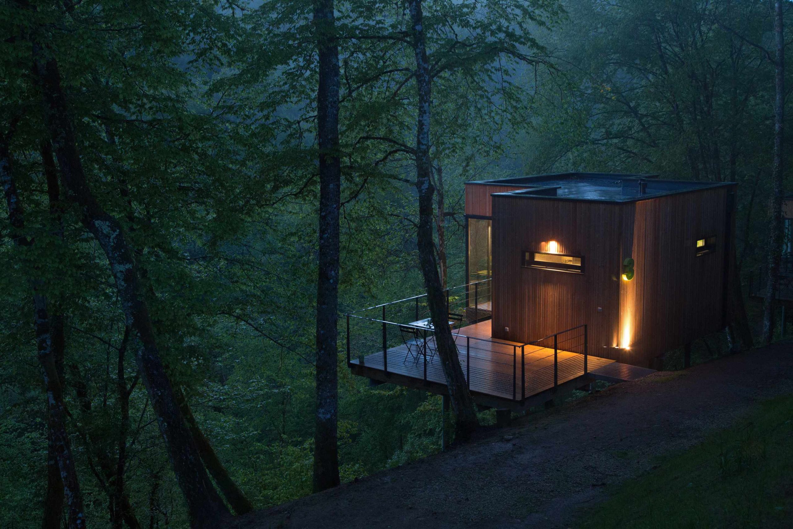 E98A7572 Cabane moderne en bois, perchée dans les arbres, illuminée au crépuscule.
