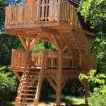 Cabane perchée en bois avec terrasse, entourée darbres verdoyants.