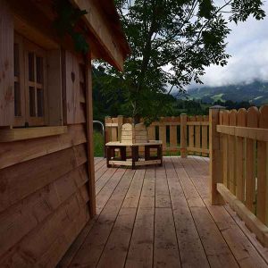 Cabane en bois avec terrasse ensoleillée et vue sur les montagnes verdoyantes.