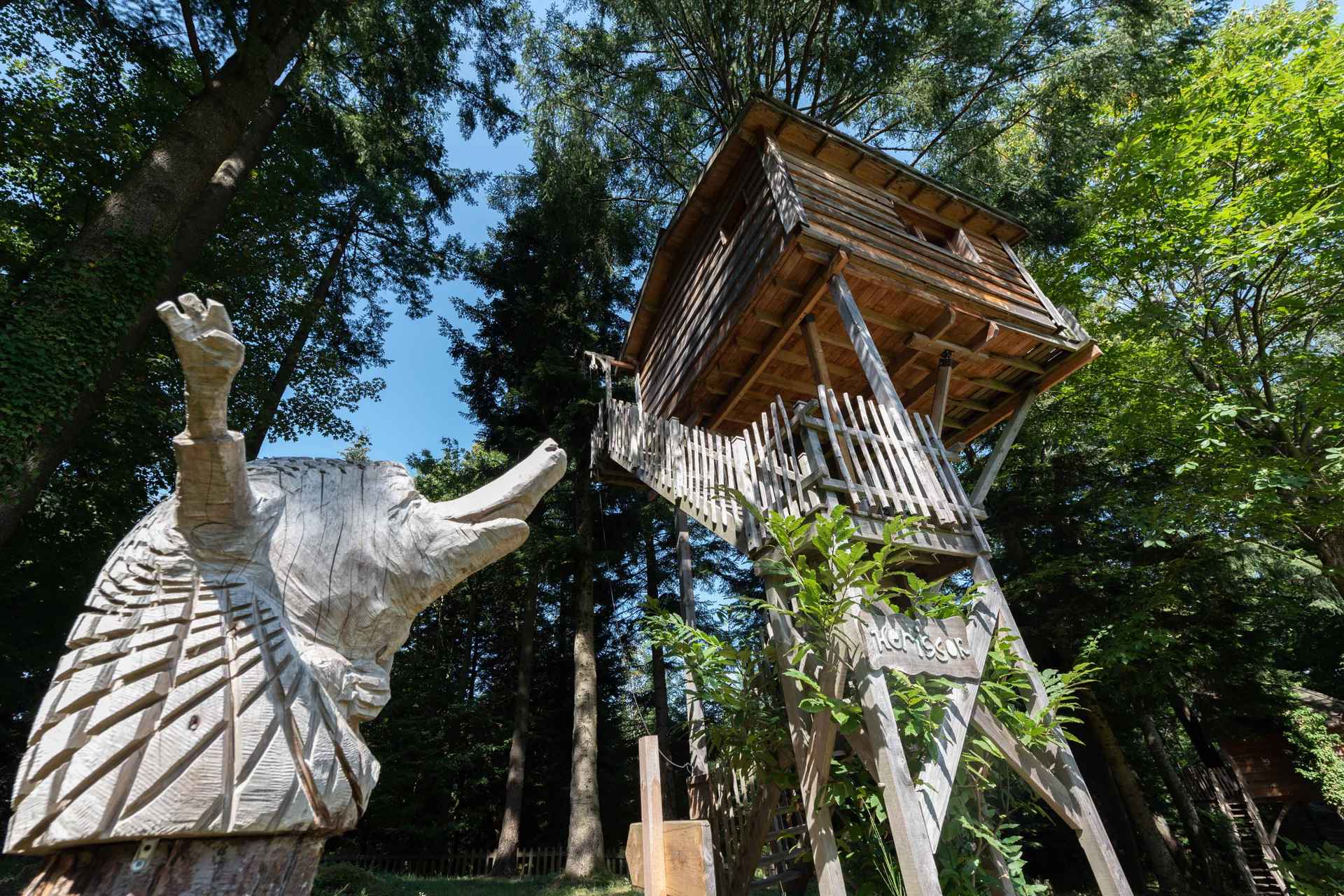 version-internet-2Herisson Cabane perchée en bois entourée darbres, avec une sculpture danimaux en bois.