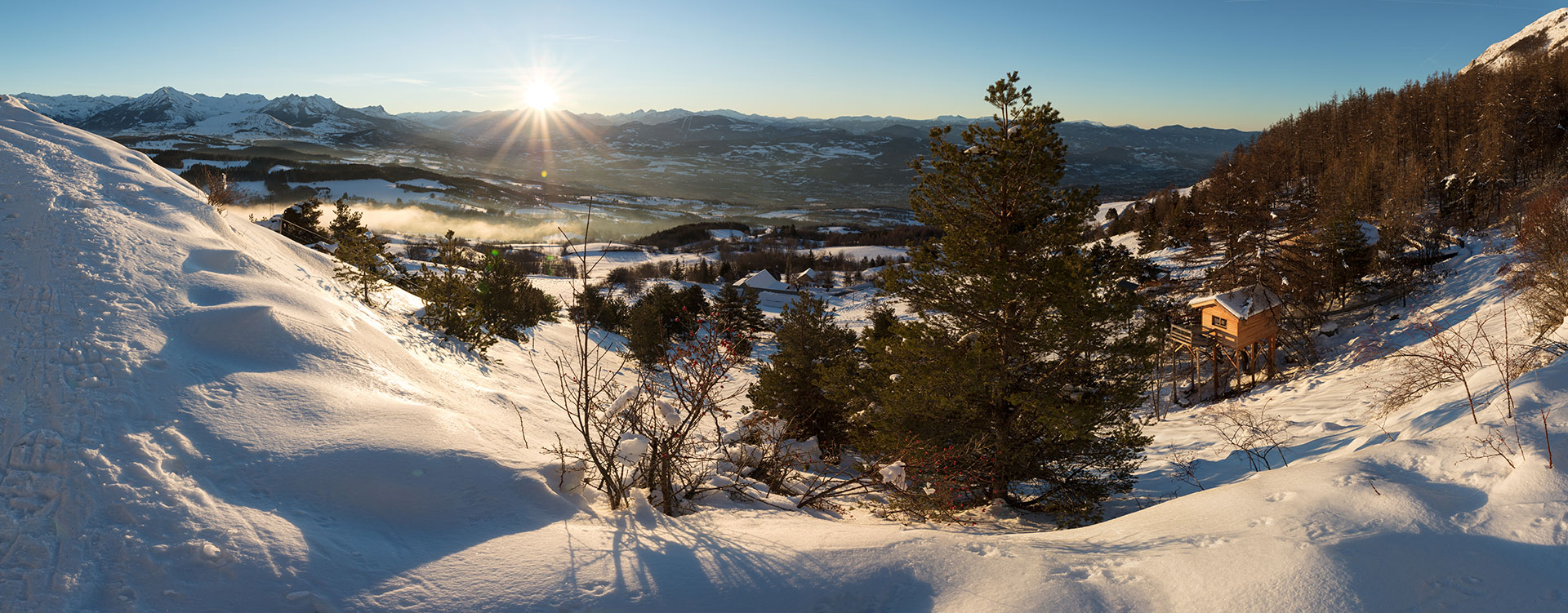 Cabane en bois perchée, vue panoramique sur les montagnes enneigées au coucher du soleil.