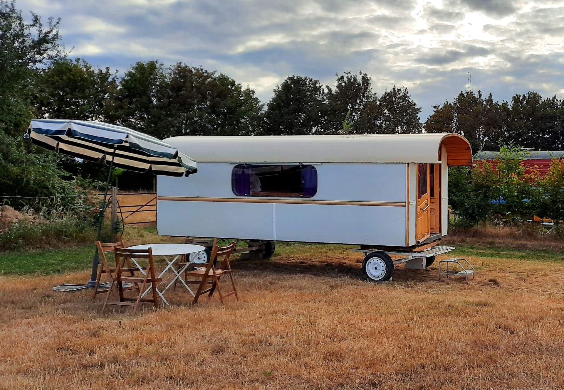 Roulotte Gitane 1 Caravane vintage sur un terrain verdoyant, avec parasol et table en bois.