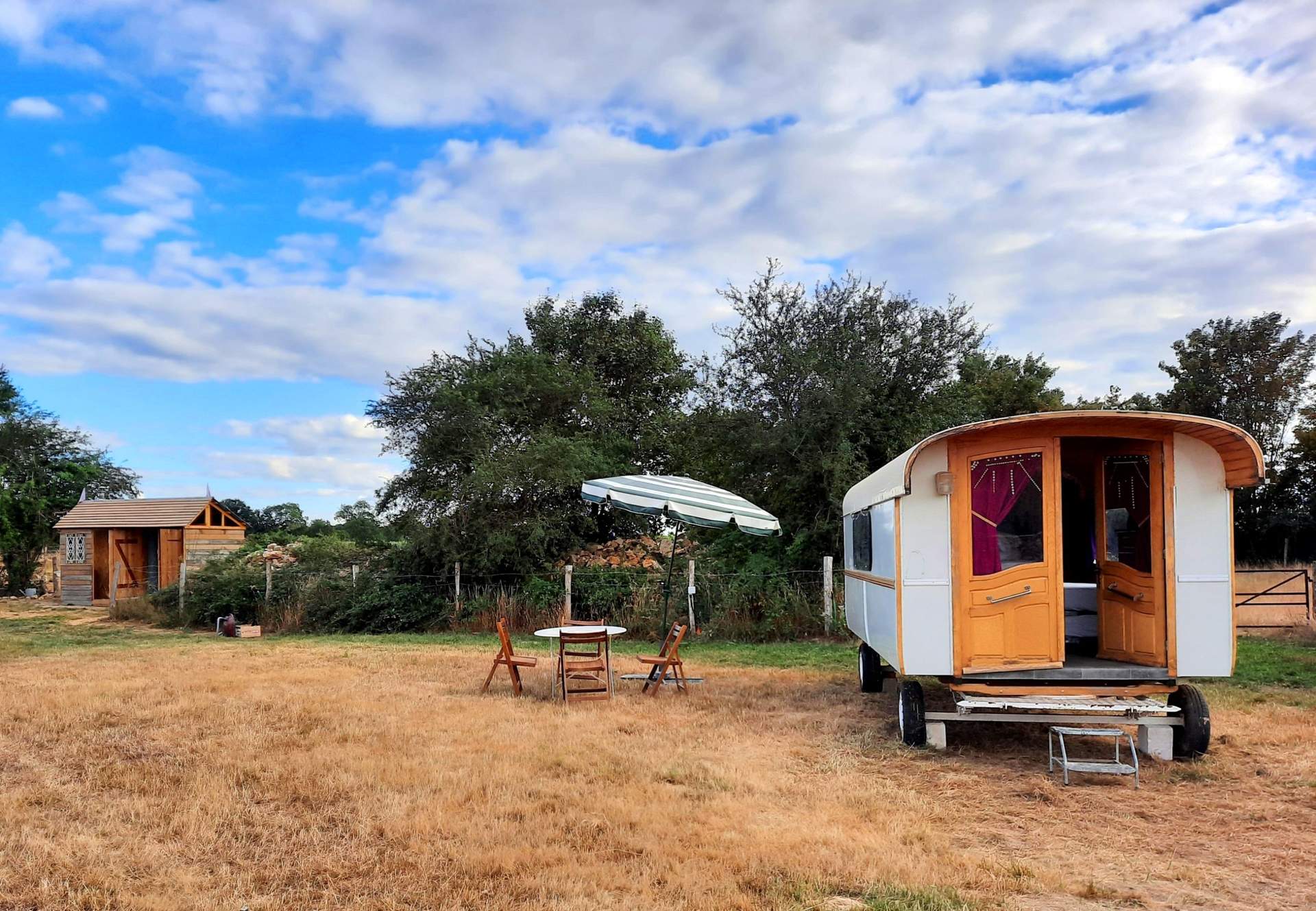 Roulotte Gitane 3 Charmante roulotte avec parasol, entourée de verdure et dun petit chalet en bois.