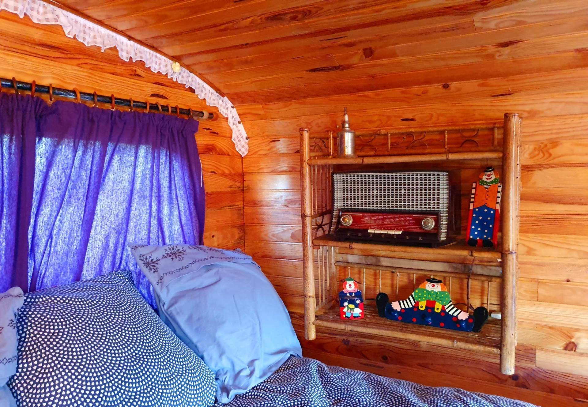 Roulotte Gitane 6 Chambre cosy dans une cabane en bois, décorée de peluches colorées et rideaux violets.