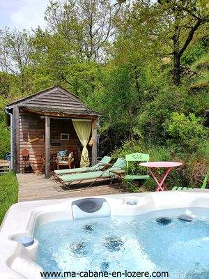 Cerf-Ma-p_tite_Cabane-en-Lozere-(4) Cabane en bois avec jacuzzi, entourée de verdure et chaises longues confortables.