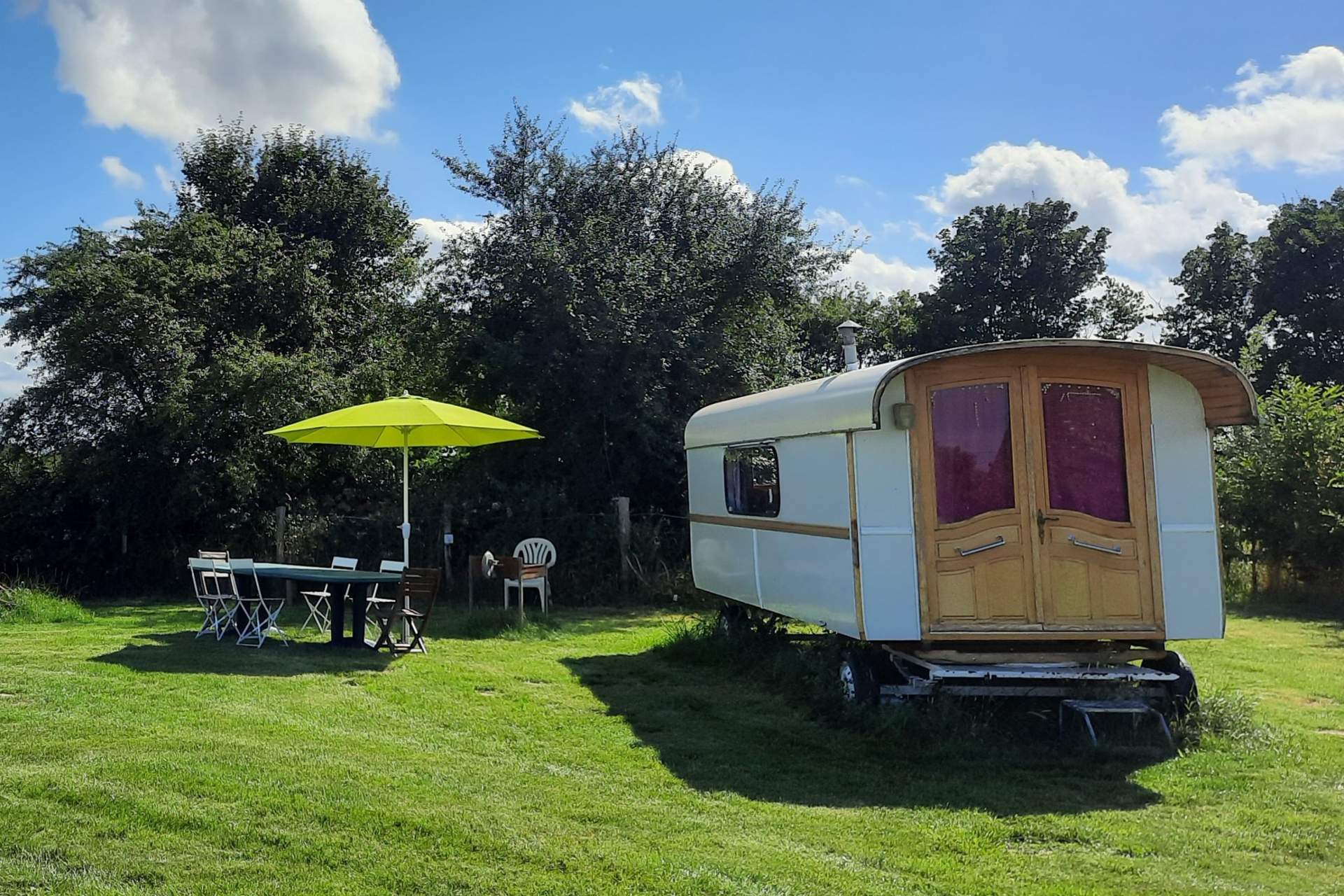 20210730_235323 Caravane vintage entourée de verdure, avec une terrasse et parasol vert.