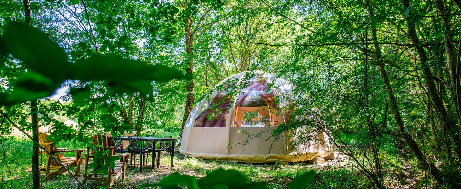 Dôme transparent en pleine nature, entouré darbres, avec mobilier de jardin.