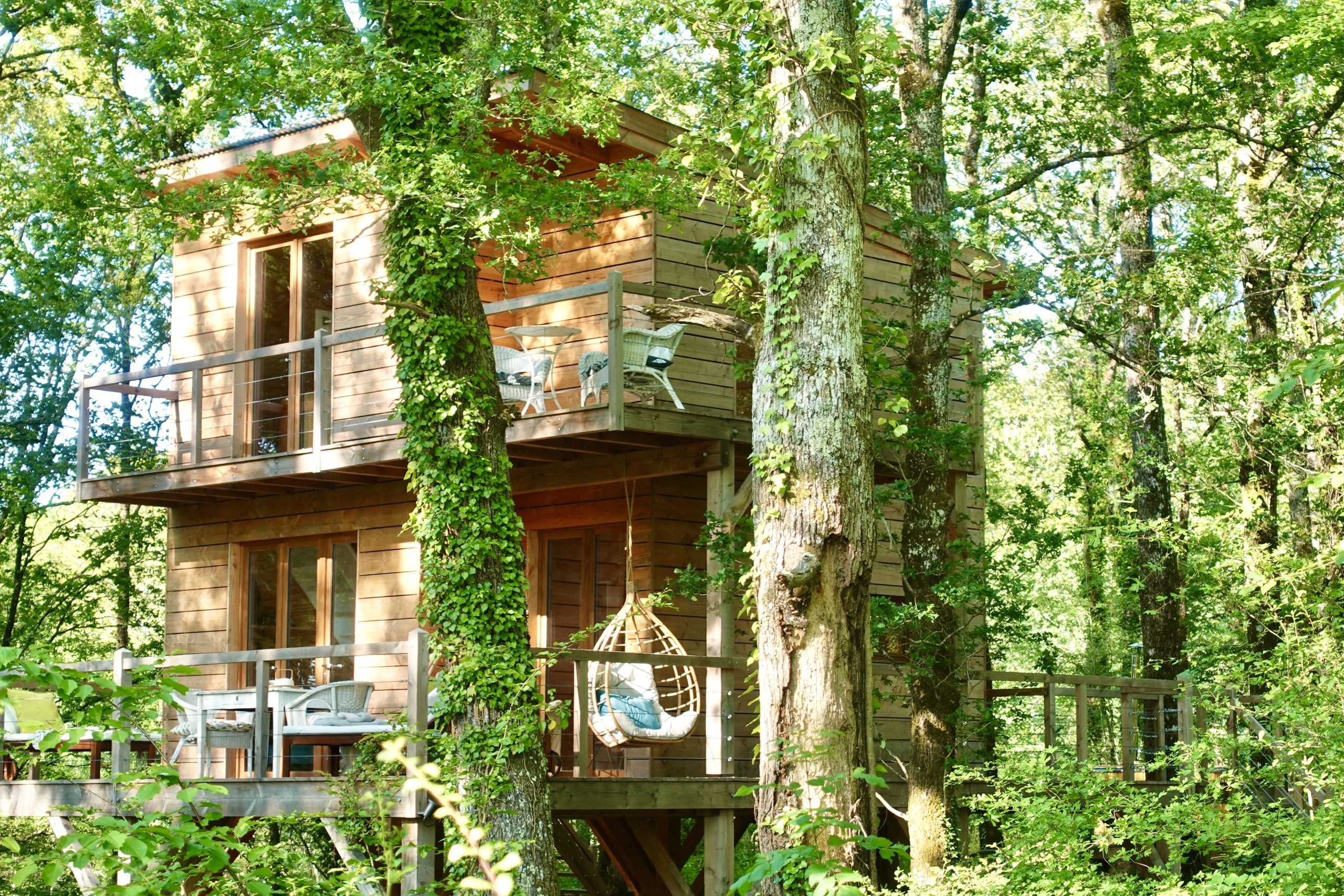Cabane perchée en bois, entourée darbres, avec balcon et chaises confortables.