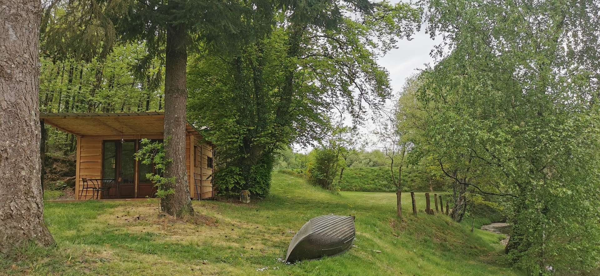 IMG_20210430_132415 Cabane en bois au bord dune rivière, entourée darbres verdoyants.