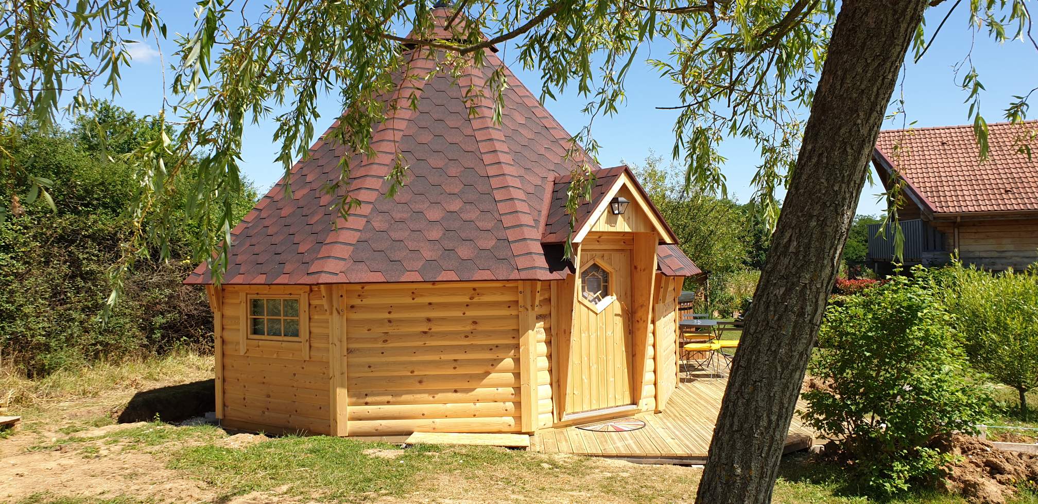 C2315A46-F72F-48C8-84AB-F45FDD1C7E44 Cabane en bois ronde avec toit en ardoise, entourée de verdure paisible.