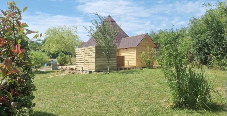 E47A5434-579D-46DA-A5DF-4FEE938B63FE Cabane en bois avec toit en pointe, entourée de verdure et dun jardin paisible.