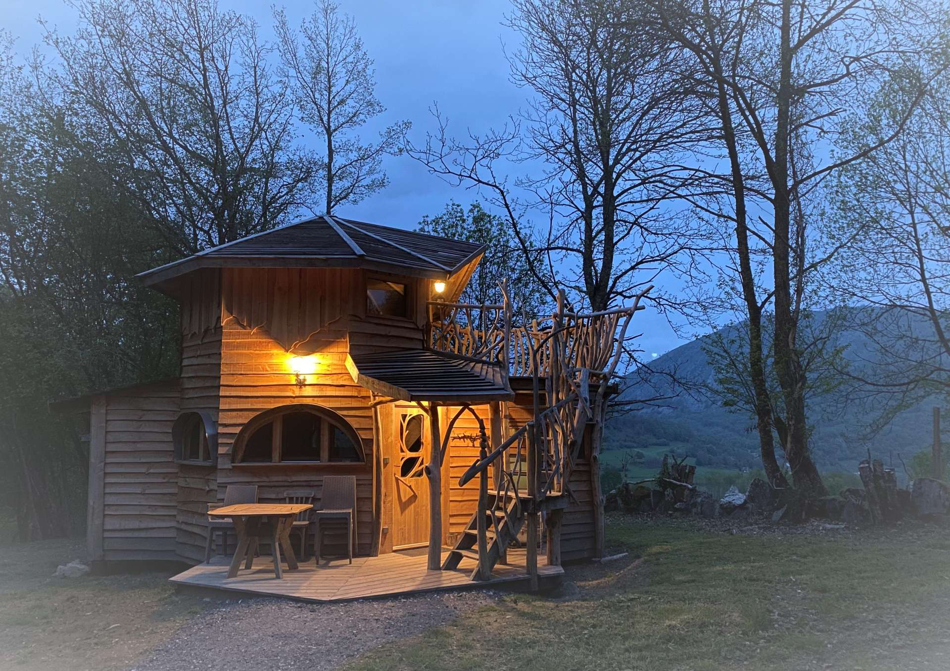 IMG_5536 Cabane perchée en bois, illuminée au crépuscule, entourée darbres verdoyants.