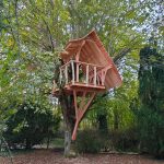 Cabane dans les arbres, perchée avec un balcon en bois, entourée de verdure.