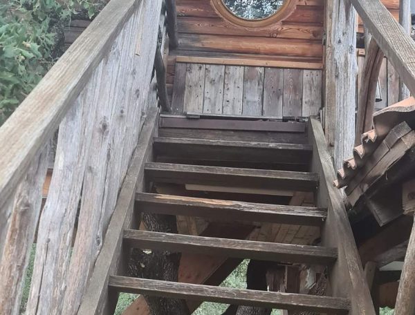 Cabane perchée en Corse, accès par un escalier en bois et fenêtre ronde.