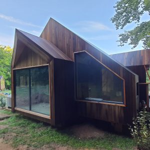 Cabane moderne en bois avec de grandes fenêtres, entourée de verdure.