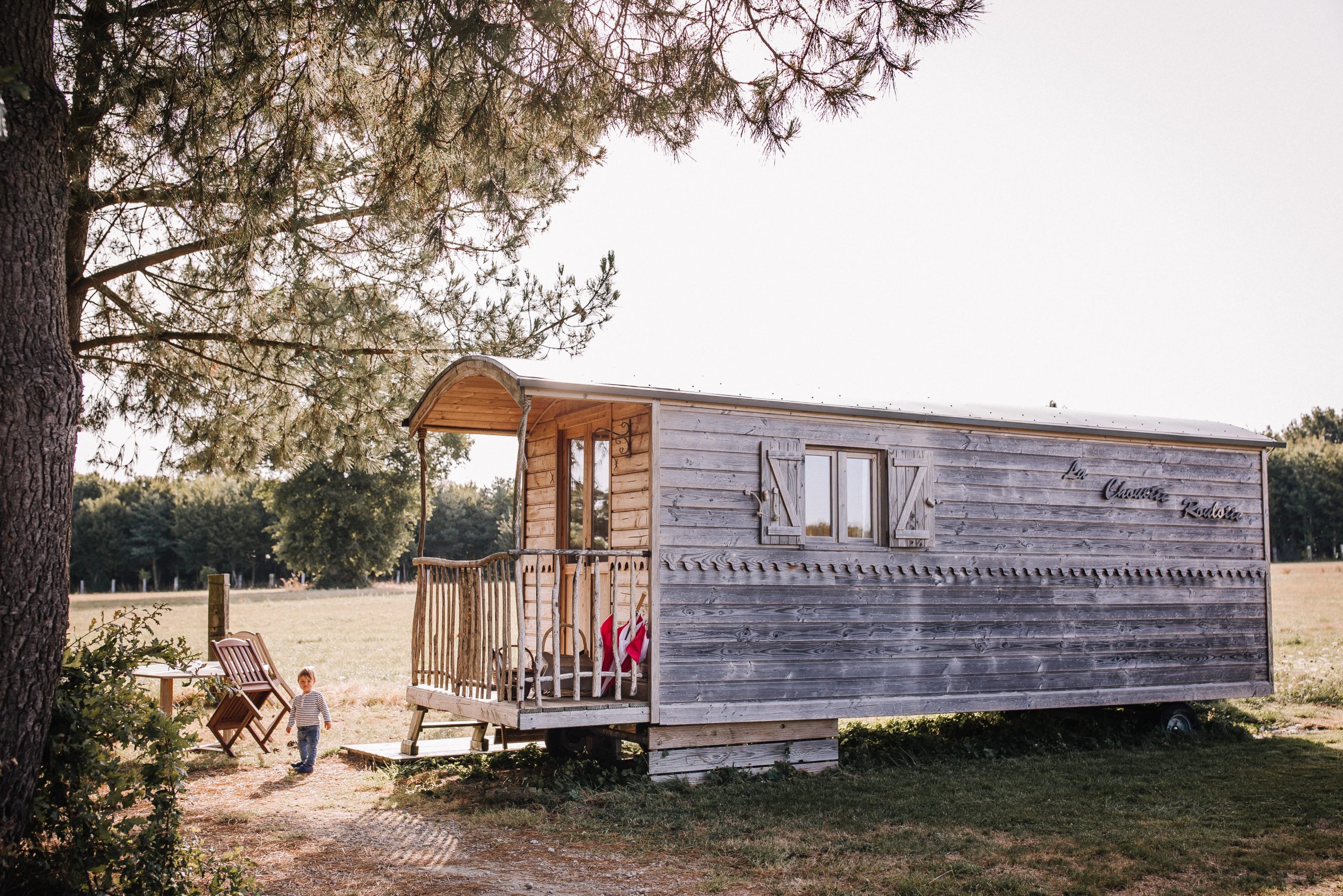 ChouetteCabane_perso_JustineB-101 Hébergement insolite en roulotte en bois, entouré de verdure et dun enfant jouant.