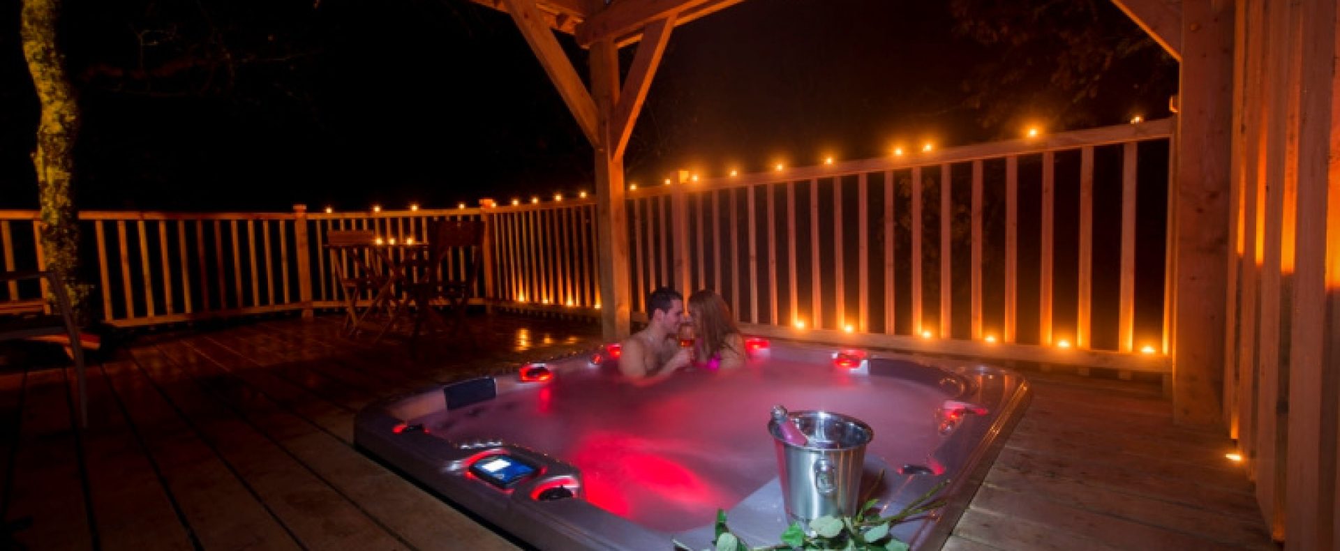 Cabane en bois avec jacuzzi romantique, éclairage doux et roses sur la terrasse.