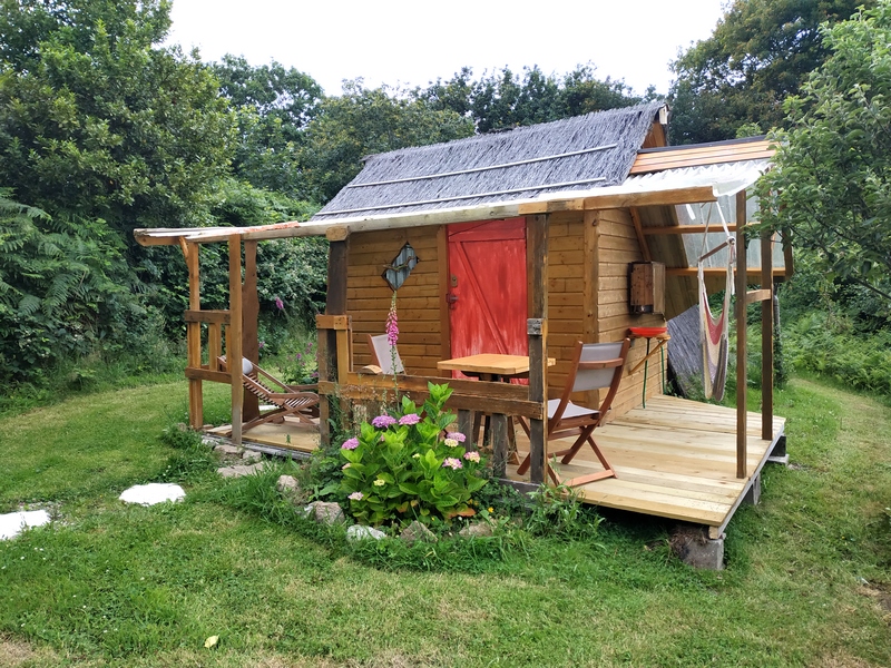 cahutte 9 Cabane en bois avec terrasse, entourée de verdure et de fleurs colorées.
