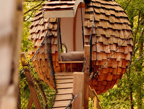 Cabane en bois en forme de sphère, perchée dans les arbres, accessible par un pont suspendu.