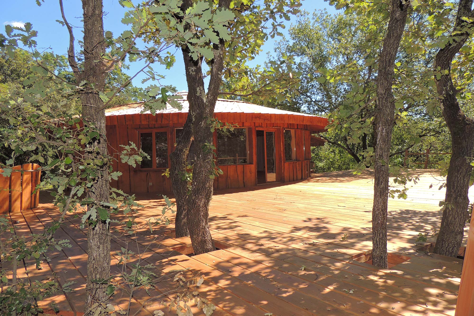 cabane-maloka-2000-exterieur5 Cabane en bois au milieu des arbres, avec une grande terrasse ensoleillée.