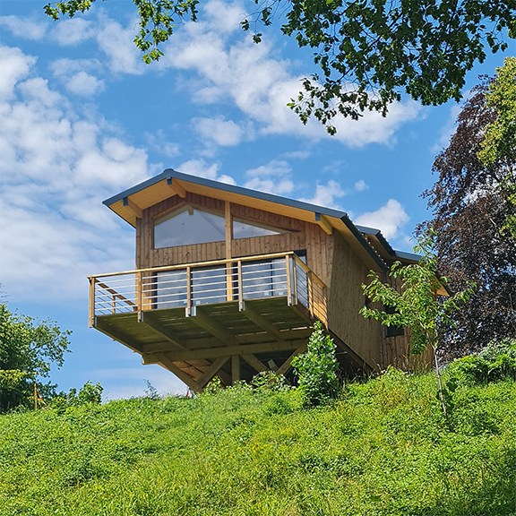 cabane_perle Cabane en bois perchée, avec terrasse ensoleillée et vue sur la nature verdoyante.