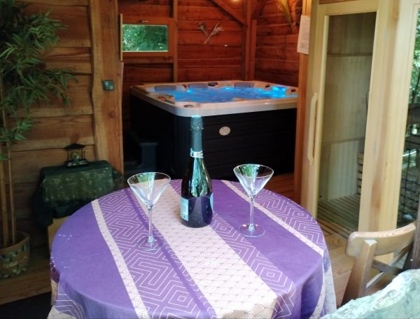 Cabane en bois avec jacuzzi, table élégante et bouteilles pour un séjour romantique.