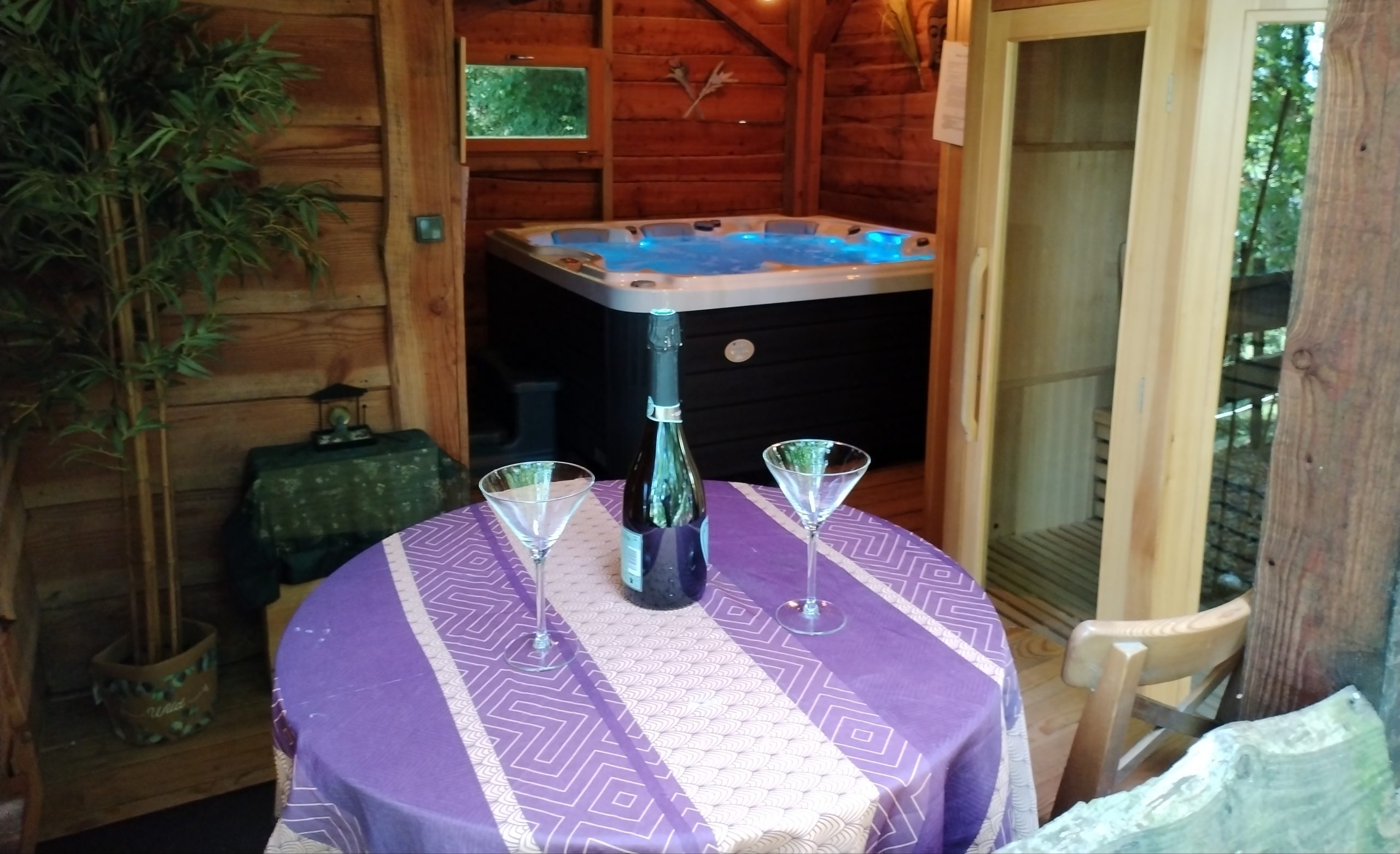 Cabane en bois avec jacuzzi, table élégante et bouteilles pour un séjour romantique.