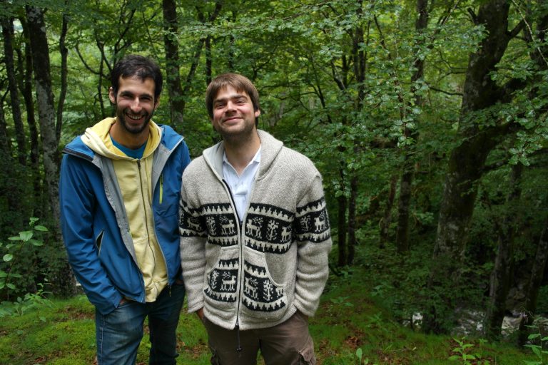 Deux amis souriants devant un chalet en bois, entouré dune forêt verdoyante.