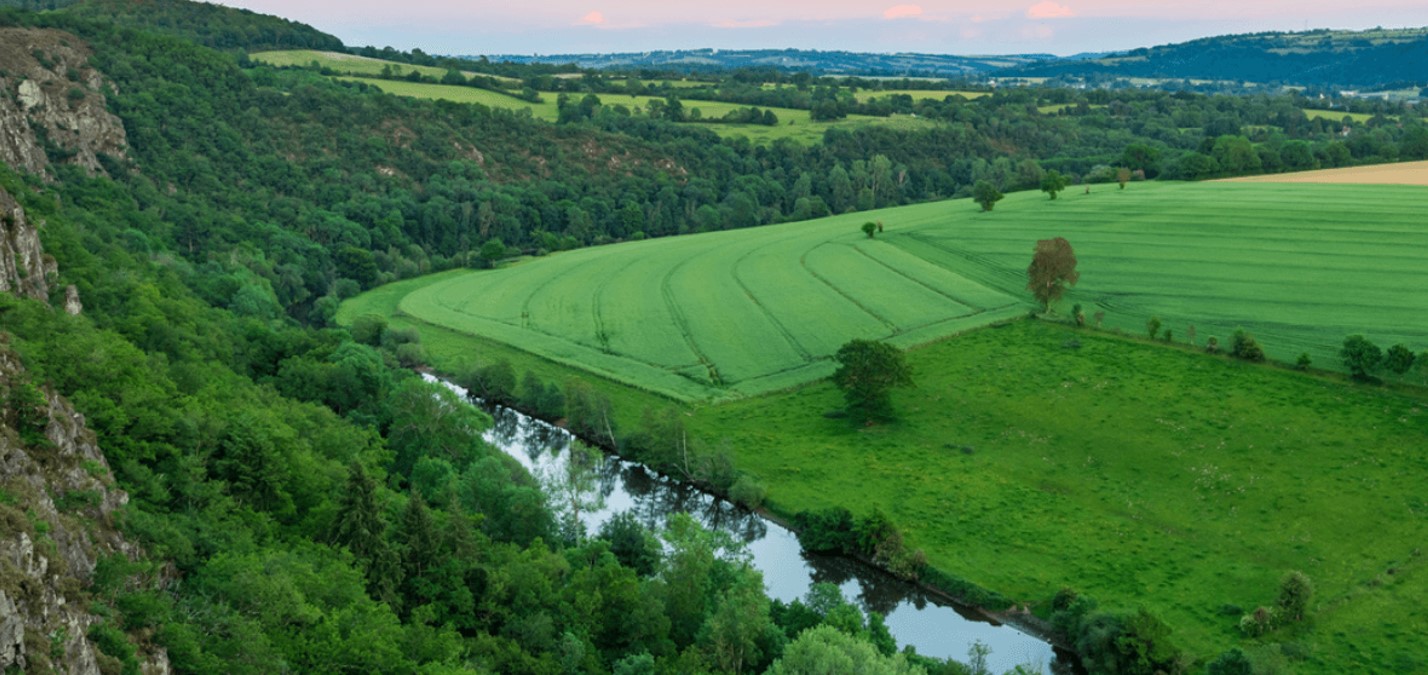 Vue panoramique dun gîte en pleine nature, entouré de champs verdoyants et dune rivière.