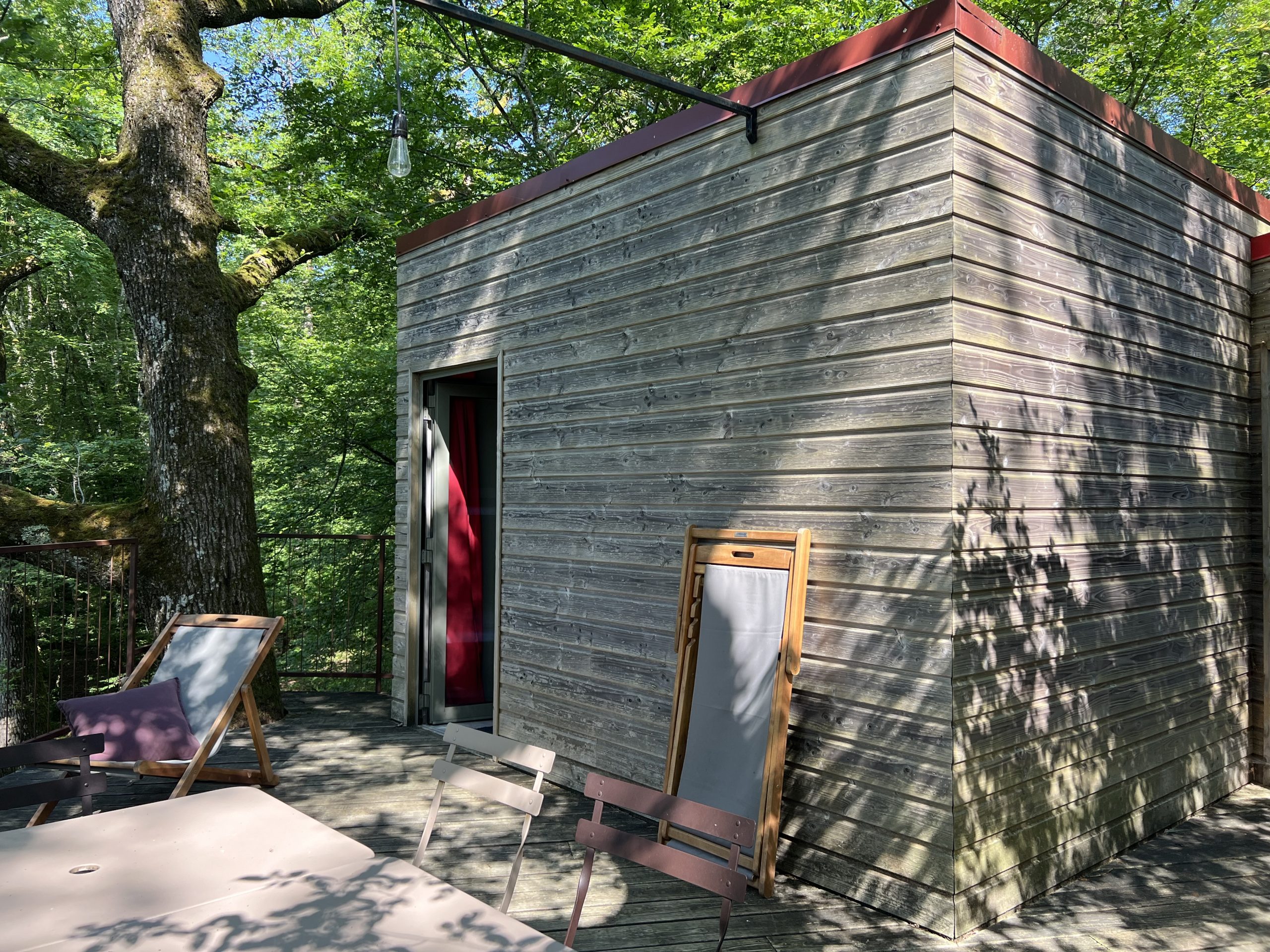 Eau10 Cabane en bois perchée, entourée darbres, avec terrasse en bois et mobilier extérieur.