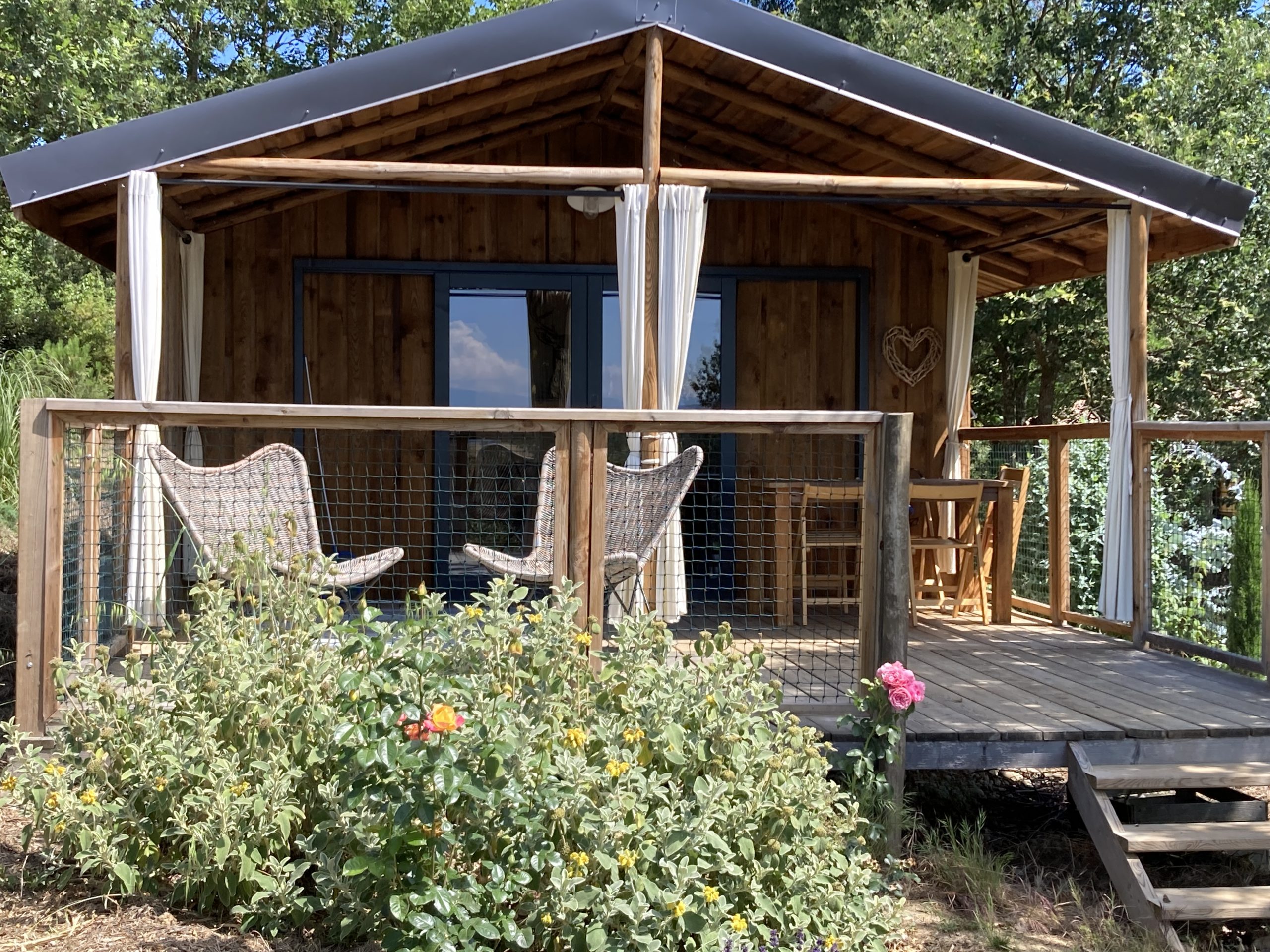 IMG_4424 Cabane en bois avec terrasse, entourée de verdure et fleurs colorées.