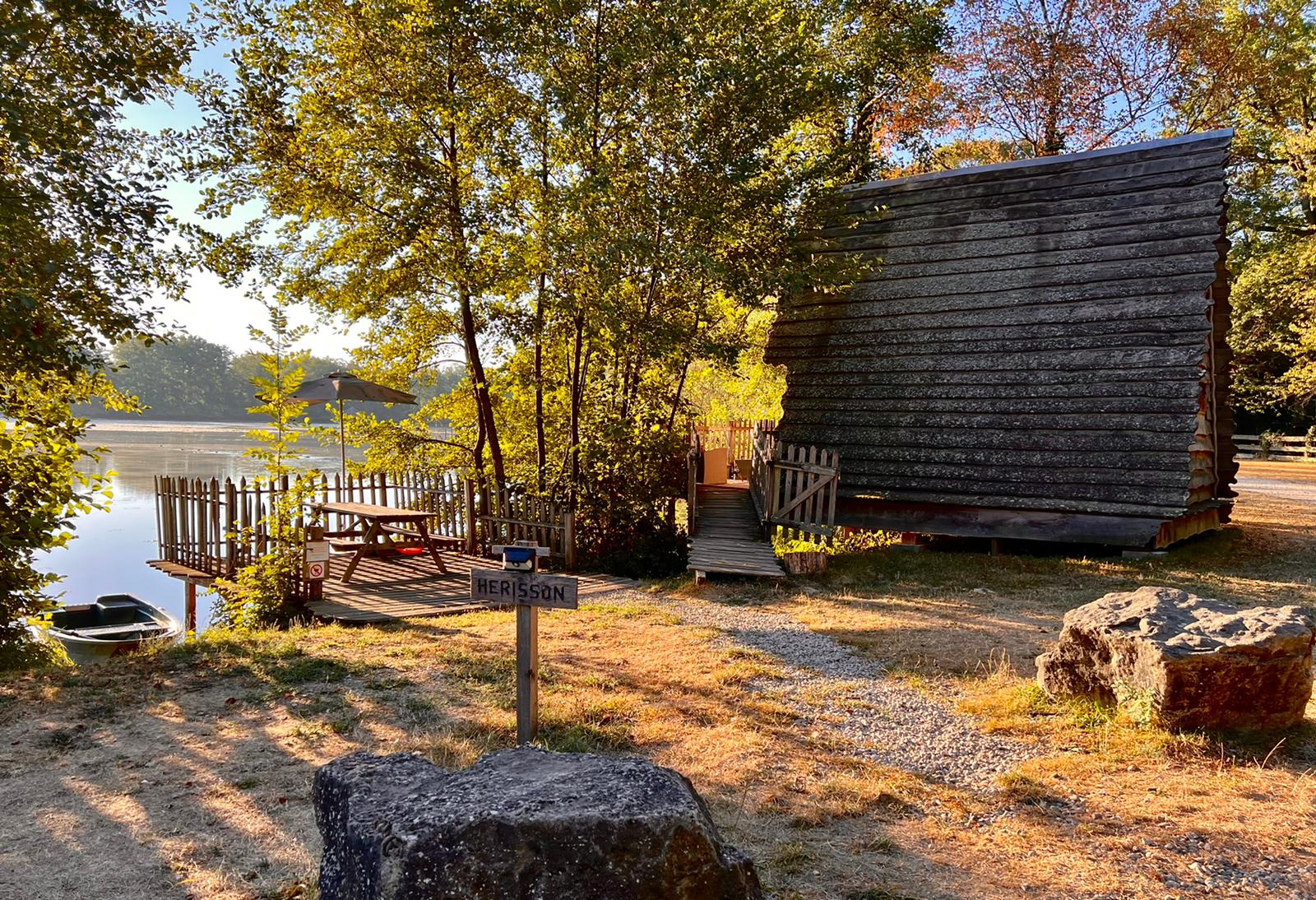 06ecf521-68bc-49f9-95fd-df78b8910fbb Cabane en bois au bord dun lac, entourée darbres et dune terrasse ensoleillée.